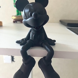 Op de afbeelding: Een zwarte Mickey Mouse-figuur die op een wit oppervlak zit. De figuur heeft grote ronde oren, een lachend gezicht en draagt een korte broek en schoenen. De benen van de figuur bungelen over de rand van het oppervlak.