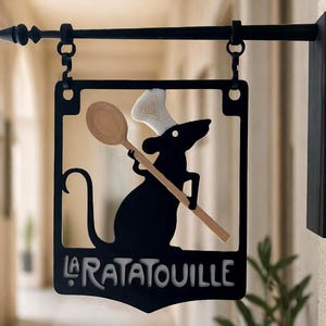 Può includere: Un'insegna da parete in metallo nero con la sagoma di un topo che indossa un cappello da chef bianco e tiene in mano un cucchiaio di legno. Il testo LA RATATOUILLE è impresso sul fondo della cornice quadrata, appesa a un'asta orizzontale.