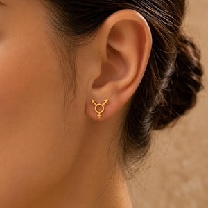 May include: A close-up of a person's ear with a pair of small, gold-colored stud earrings. Each earring features the transgender symbol. The earrings are simple in design and have a polished finish. The background is a soft, neutral color.