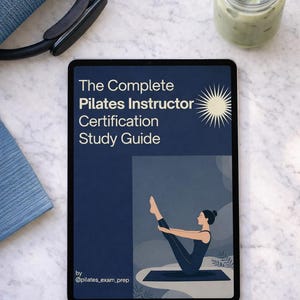 May include: A tablet displays "The Complete Pilates Instructor Certification Study Guide" with a sunburst graphic and a Pilates illustration. A Pilates ring, a blue mat, and a green drink in a glass are also visible.