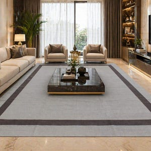 Op de afbeelding: Een lichtgrijs rechthoekig vloerkleed met een donkerbruine rand. Het vloerkleed bevindt zich in een woonkamer met een bank, stoelen en een salontafel. De kamer heeft grote ramen en een modern design.