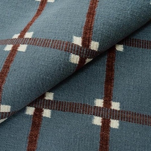 Può includere: Primo piano di un tessuto blu con un motivo a quadri. Il design presenta linee verticali e orizzontali in marrone scuro, che si intersecano con accenti quadrati bianchi. Il tessuto è piegato, mostrando la sua trama.