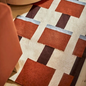 May include: A rectangular area rug featuring a geometric pattern of terracotta orange, dark brown, and light cream blocks. The design includes horizontal light blue accents along the top edges of the orange rectangles, creating a structured layout.