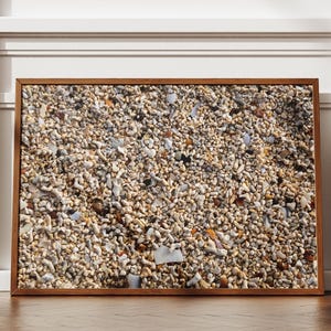 May include: A framed print featuring a close-up view of a beach composed of small, light-colored pebbles, shells, and coral fragments. The artwork is encased in a brown wooden frame, creating a natural and textured aesthetic. The print is displayed against a white wall.