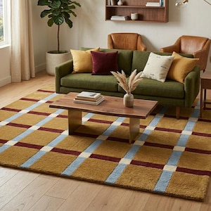 Puede incluir: Una alfombra rectangular grande con un patrón de cuadrícula en amarillo mostaza, azul claro y burdeos intenso. La alfombra tiene una superficie texturizada y copetuda, cubriendo gran parte del suelo de madera clara en la sala de estar.