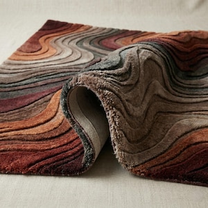 May include: A rectangular area rug featuring a textured, wavy pattern in shades of deep red, burnt orange, brown, and muted gray. The rug is partially folded, revealing a light-colored, woven fabric backing on the underside of the corner.