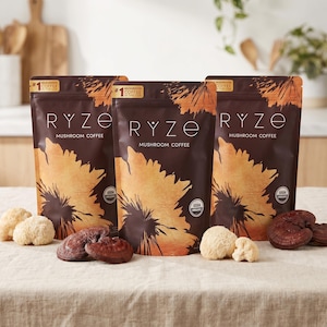 Puede incluir: Tres bolsas marrones de caf&eacute; de hongos RYZE con un dise&ntilde;o de girasol. El texto "Mushroom Coffee" y "#1 Coffee in the USA" son visibles. Varias setas est&aacute;n dispuestas frente a las bolsas, incluyendo melena de le&oacute;n y reishi.