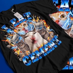 May include: A black t-shirt featuring a graphic print of multiple cats, bags labelled CEMENT, blue lightning bolts, and fire. The text I EAT CEMENT is printed in large, bold, blue letters across the design. A white price tag is attached to the collar.