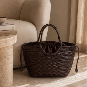 May include: A dark brown woven tote bag featuring a structured silhouette with two rounded handles and a drawstring closure. The exterior displays a consistent braided pattern, while the interior is lined with a dark brown fabric.