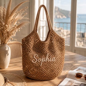 Può includere: Una borsa tote all'uncinetto color marrone chiaro con un design a rete aperta e due tracolle. La parte anteriore della borsa mostra il nome Sophia ricamato in corsivo bianco al centro del materiale intrecciato.