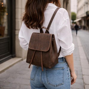 Puede incluir: Una mochila de cuero marrón con cierre de solapa y detalles de cordón. La mochila tiene correas ajustables. La persona lleva una camisa blanca y vaqueros azules.