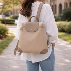 May include: A beige and taupe backpack with gold-colored zippers and hardware. The backpack has a flap closure and a top handle. The straps are adjustable. The bag is made of a smooth material and is being worn.