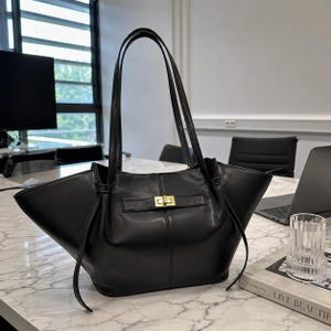 May include: A black leather tote bag with long handles. The bag features a gold-colored clasp and a structured design. The bag is sitting on a marble table.