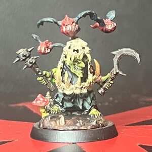 May include: A miniature fantasy figure of a green-skinned creature with a bone-like headdress, holding a spiked mace and a curved blade. It has three floating, red-mouthed creatures above its head and stands on a detailed base with a mushroom.