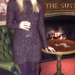 May include: A woman wearing a sparkly, dark purple dress with long sleeves and a round neckline. She is holding a glass of liquid in her right hand. She is standing in front of a fireplace with a sign that reads "The Sussex" above it.
