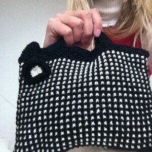 Könnte beinhalten: Eine schwarze und weiße gehäkelte Handtasche mit einem weißen Blumen-Detail. Die Tasche hat ein strukturiertes, gitterartiges Muster.