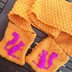 May include: Two knitted orange scarves with pink cat designs. The scarves are made with a textured stitch and have a fringed edge.