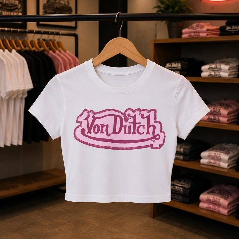 May include: A white short-sleeved crop top featuring a prominent pink Von Dutch logo printed across the chest. The garment has a round neckline and is displayed on a wooden hanger, showcasing a clean design with a bold, stylized brand graphic.