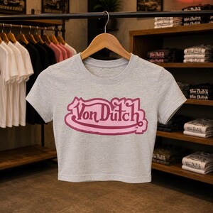 May include: A heather gray short-sleeved crop top featuring a prominent pink Von Dutch logo printed on the center chest. The garment has a round neckline and is displayed on a wooden hanger, showcasing a simple design with a bold brand graphic.