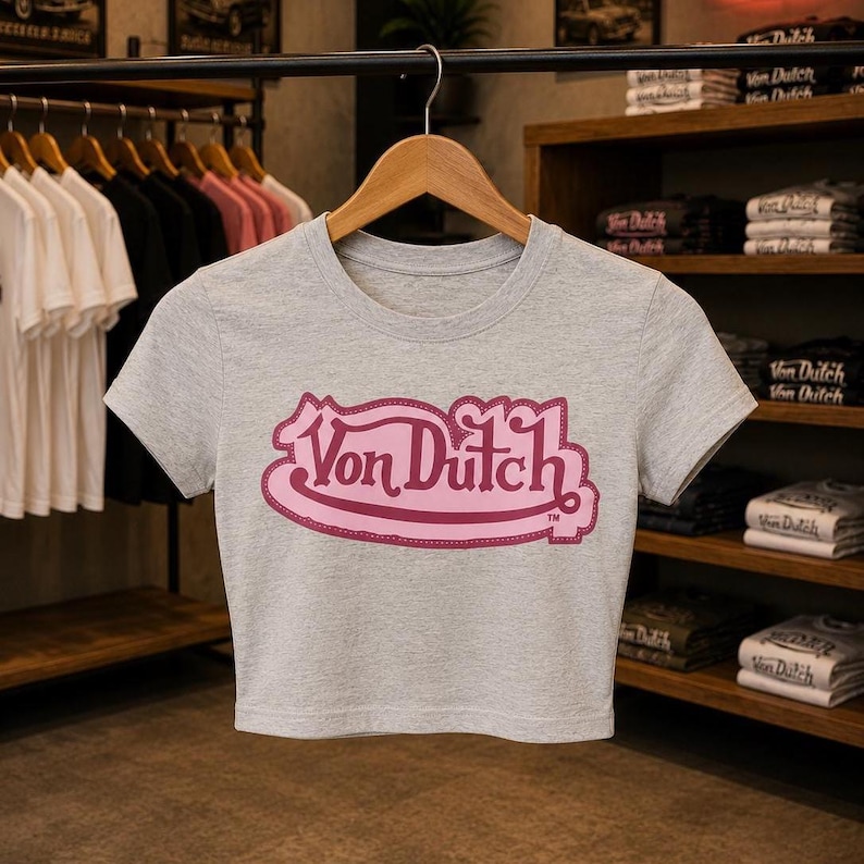 May include: A heather gray short-sleeved crop top featuring a prominent pink Von Dutch logo printed on the center chest. The garment has a round neckline and is displayed on a wooden hanger, showcasing a simple design with a bold brand graphic.