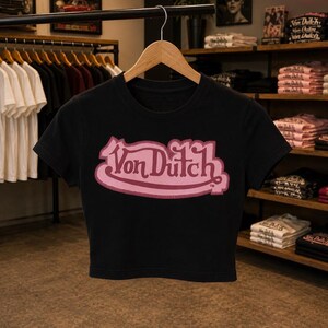 May include: A black short-sleeved crop top featuring a prominent pink Von Dutch logo printed across the center of the chest. The garment has a round neckline and is displayed on a wooden hanger, showcasing a clean, fitted silhouette and bold text.