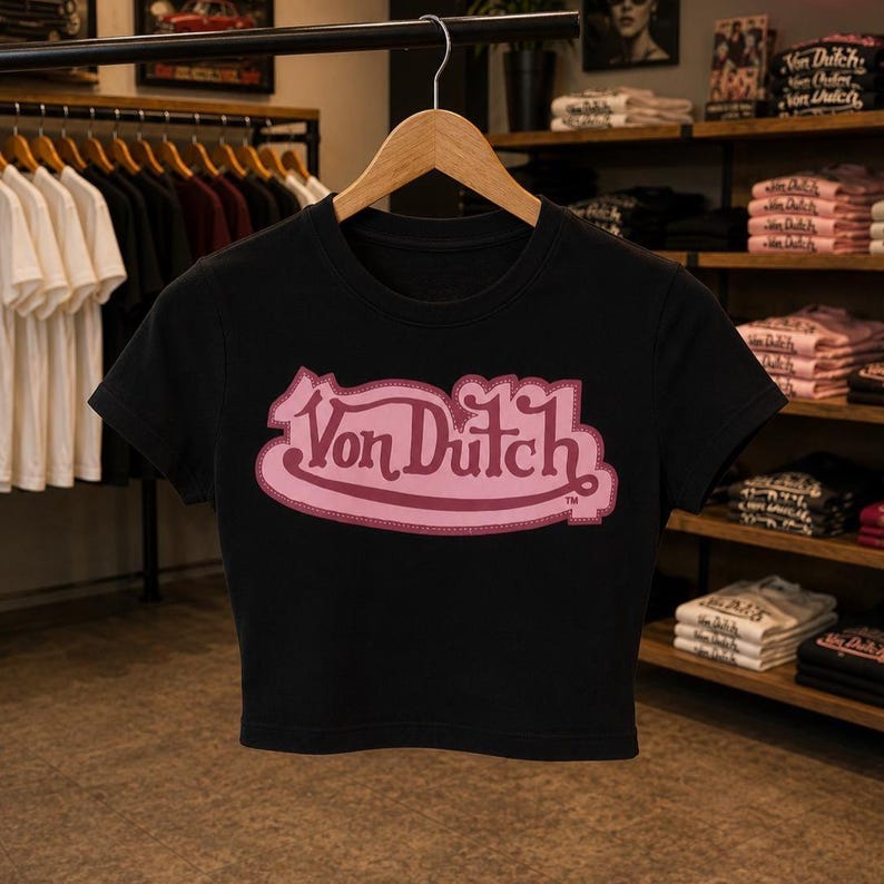 May include: A black short-sleeved crop top featuring a prominent pink Von Dutch logo printed across the center of the chest. The garment has a round neckline and is displayed on a wooden hanger, showcasing a clean, fitted silhouette and bold text.