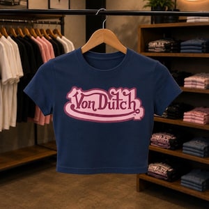May include: A navy blue short-sleeved crop top featuring the text Von Dutch in a stylized pink font with a dark pink outline centered on the chest. The garment is displayed on a wooden hanger against a background of clothing racks and shelves.