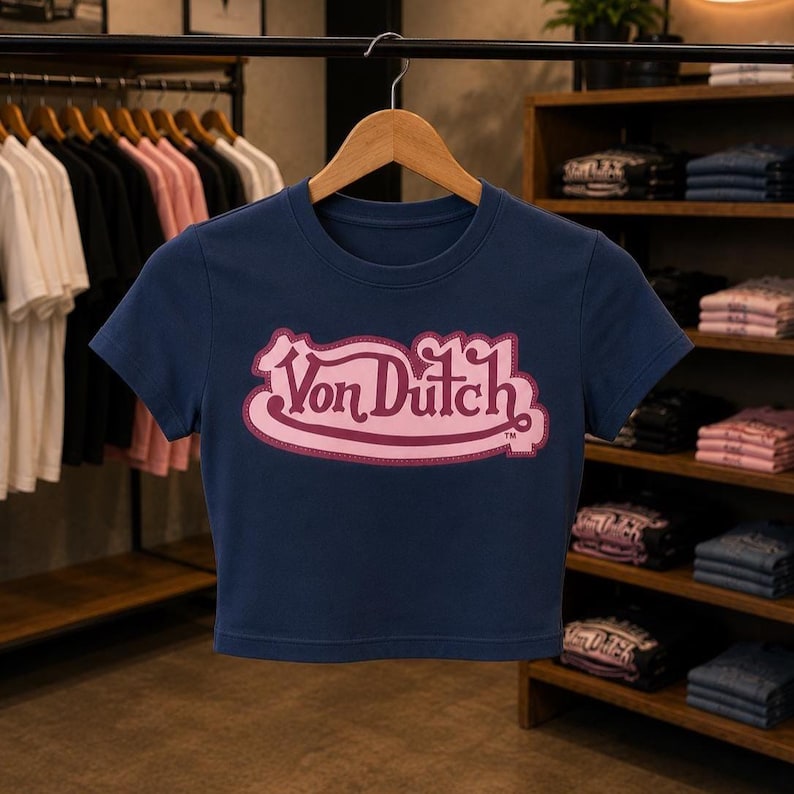 May include: A navy blue short-sleeved crop top featuring the text Von Dutch in a stylized pink font with a dark pink outline centered on the chest. The garment is displayed on a wooden hanger against a background of clothing racks and shelves.