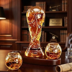 May include: A set of crystal glassware, including a trophy-shaped decanter and two spherical glasses, all filled with amber liquid. The decanter is the shape of a trophy. The glasses have a soccer ball design. The set is displayed on a wooden base.