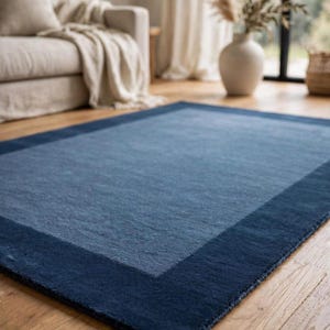 Könnte beinhalten: Ein rechteckiger Teppich mit einer hellblauen Mittelfläche, die von einem breiten, dunkel marineblauen Rand umgeben ist. Der Teppich hat eine kurzflorige Textur und liegt flach auf einem hellen Holzboden, was ein klares, zweifarbiges Design zeigt.