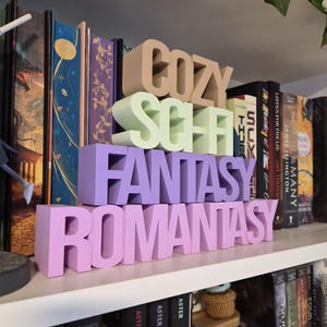 May include: A white bookshelf displaying 3D-printed words in various colours: "COZY", "SCHELF", "FANTASY", and "ROMANTASY". The letters are in beige, green, purple, and pink. Books are arranged behind the words, indicating a literary theme.