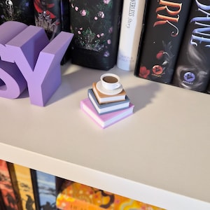 Pode incluir: Uma pilha de livros em miniatura em tons de rosa, cinza e bege, encimada por uma pequena chávena e pires brancos. As letras "SY" estão em letras grandes de lavanda. Livros com capas escuras ao fundo.