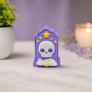 May include: A purple, tombstone-shaped trinket box with a white skull and yellow stars. The box has a cloud design at the base. The background includes a small plant, a lit candle, and lavender sprigs.