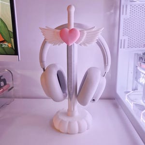 May include: A white headphone stand with a sword design, adorned with a pink heart and angel wings. It supports a pair of white headphones. The stand's base has a scalloped design. The background is a soft, light purple.