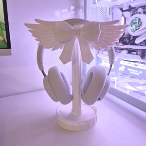 Peut inclure: Un support de casque blanc composé d'un pilier central avec une base circulaire et une pièce décorative supérieure en forme de nœud avec des ailes déployées. Le support maintient un casque audio blanc, présentant une teinte uniforme.