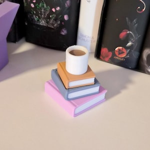 May include: A stack of three miniature books in pink, blue, and tan, topped with a small white cup. The books are arranged in a pyramid shape. The background includes other books and a purple geometric shape.