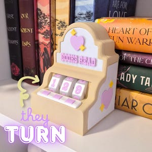 Può includere: Un contatore di libri beige e bianco in stile slot machine, con le parole "BOOKS READ" e i numeri 129. Presenta cuori rosa e stelle gialle. Libri sullo sfondo.