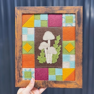 May include: A framed felt wall art piece featuring a central dark brown rectangle with three light gray mushrooms and green foliage. The border consists of a colorful patchwork quilt pattern with squares and triangles in orange, yellow, magenta, and light blue.