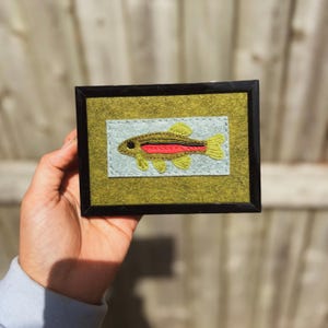 May include: A rectangular framed artwork featuring a felt applique of a fish. The fish is embroidered with yellow fins, a red lateral stripe, and a black eye, set against a light blue felt rectangle on a dark olive green felt background.