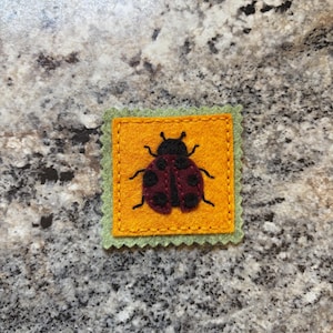 May include: A handmade felt ladybug applique. The ladybug is red with black spots and a black head, centered on a yellow square with a green scalloped border. The applique is likely for crafts or embellishment.