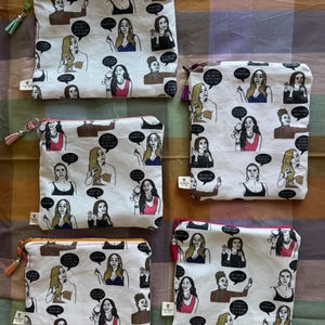 May include: Five white fabric zipper pouches featuring a repeating graphic print of various women in different outfits with black speech bubbles containing text. Each pouch has a colored zipper and a matching tassel attached to the zipper pull.