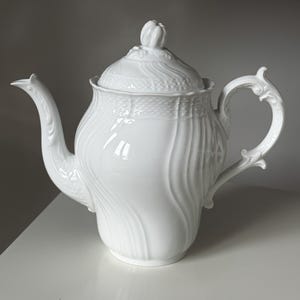 May include: A white porcelain teapot with a lid and a curved handle. The teapot features a floral design on the lid and a curved spout. The teapot is on a white surface. The teapot is made of porcelain.