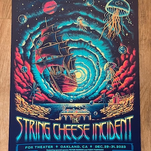 May include: A psychedelic poster for The String Cheese Incident concert at the Fox Theater in Oakland, California. The poster features a ship sailing through a swirling galaxy with jellyfish and stars. The text "The String Cheese Incident" is in a colorful, stylized font.