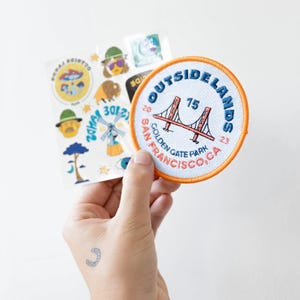 May include: A circular embroidered patch with an orange border featuring a red Golden Gate Bridge illustration, blue text reading OUTSIDELANDS, and red text reading SAN FRANCISCO, CA. Beside it is a sheet of stickers with various colorful designs.