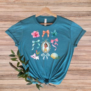 May include: Teal t-shirt with a vibrant graphic design. The design incorporates a portrait of a woman, bows, flowers, a seashell, and cartoon characters. The shirt is displayed on a wooden hanger against a wooden backdrop.