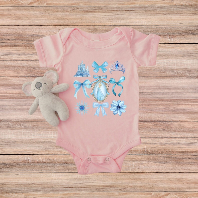 May include: A pale pink baby bodysuit decorated with blue watercolour illustrations of a castle, bows, a tiara, flowers, and a princess portrait. A plush koala toy is next to the bodysuit.