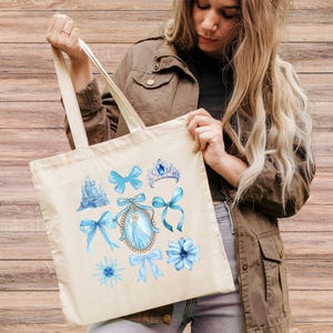 May include: A cream-coloured tote bag displaying watercolour illustrations of a blue castle, bows, a tiara, a snowflake, a flower, and a princess portrait. The bag has cream handles and is made of a durable fabric.