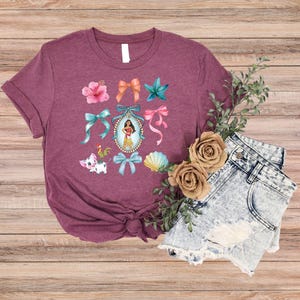 May include: A maroon t-shirt with a vibrant graphic of bows, flowers, and a cartoon character in a frame. The design includes a pig, a rooster, and a seashell. The shirt is styled with denim shorts.