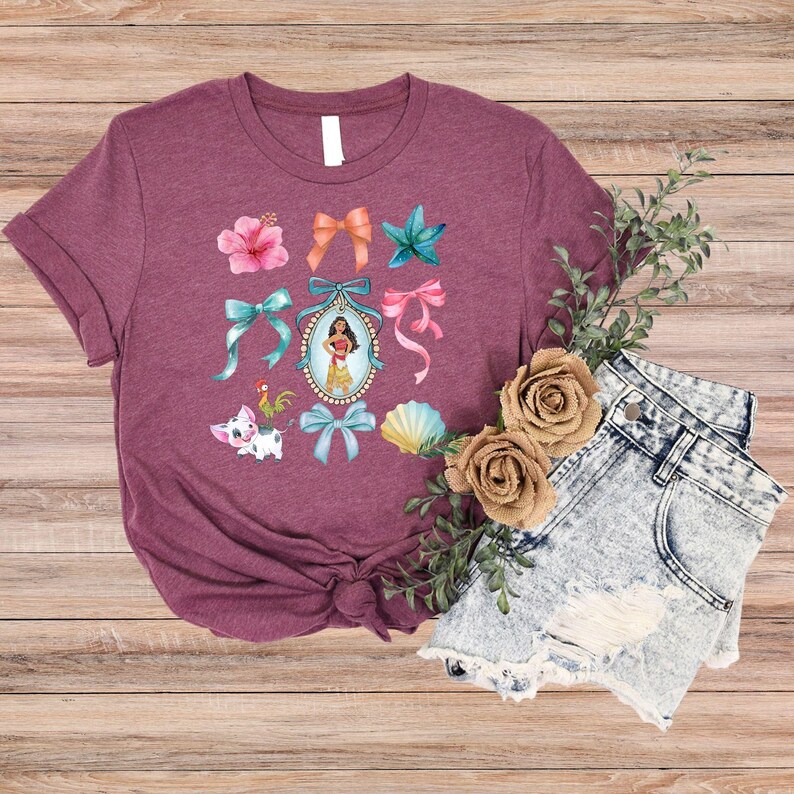 May include: A maroon t-shirt with a vibrant graphic of bows, flowers, and a cartoon character in a frame. The design includes a pig, a rooster, and a seashell. The shirt is styled with denim shorts.