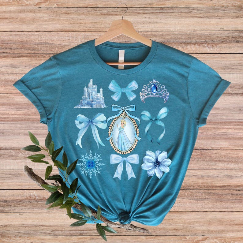 May include: Teal t-shirt with a collection of blue watercolour illustrations. The design includes a castle, bows, a tiara, a snowflake, a flower, and a portrait of a woman in a blue dress. The shirt is hanging on a wooden hanger.